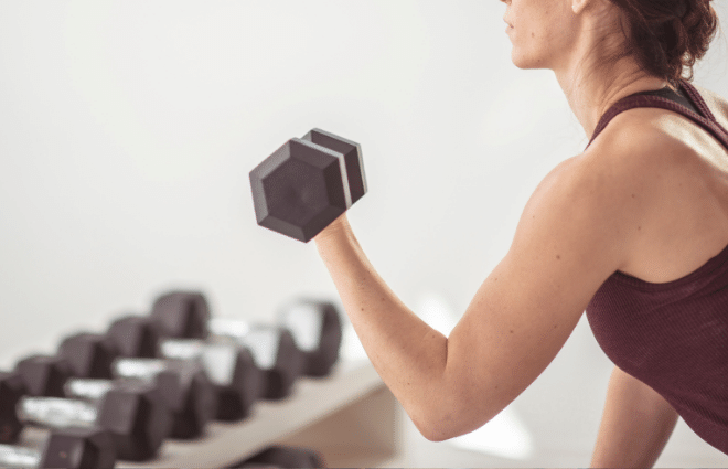 Woman lifting weights in a biceps curl with other weights on weight rack in the background. 